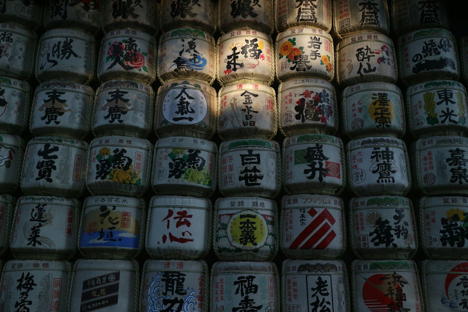 A stacked wall of decorative sake barrels wrapped in straw, each printed with a brewery's name