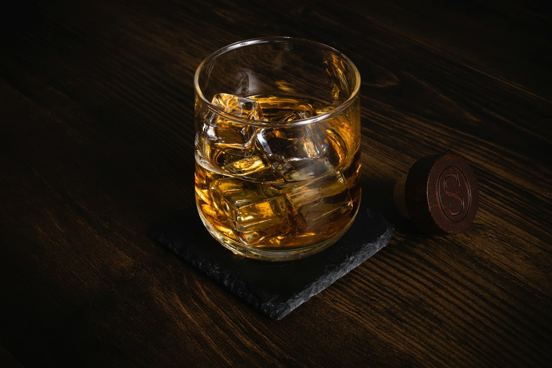 A tumbler of whisky over ice on a dark wooden table